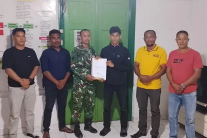 Six men pose indoors as a soldier in camouflage hands a document to a young man in black; others watch the handover.